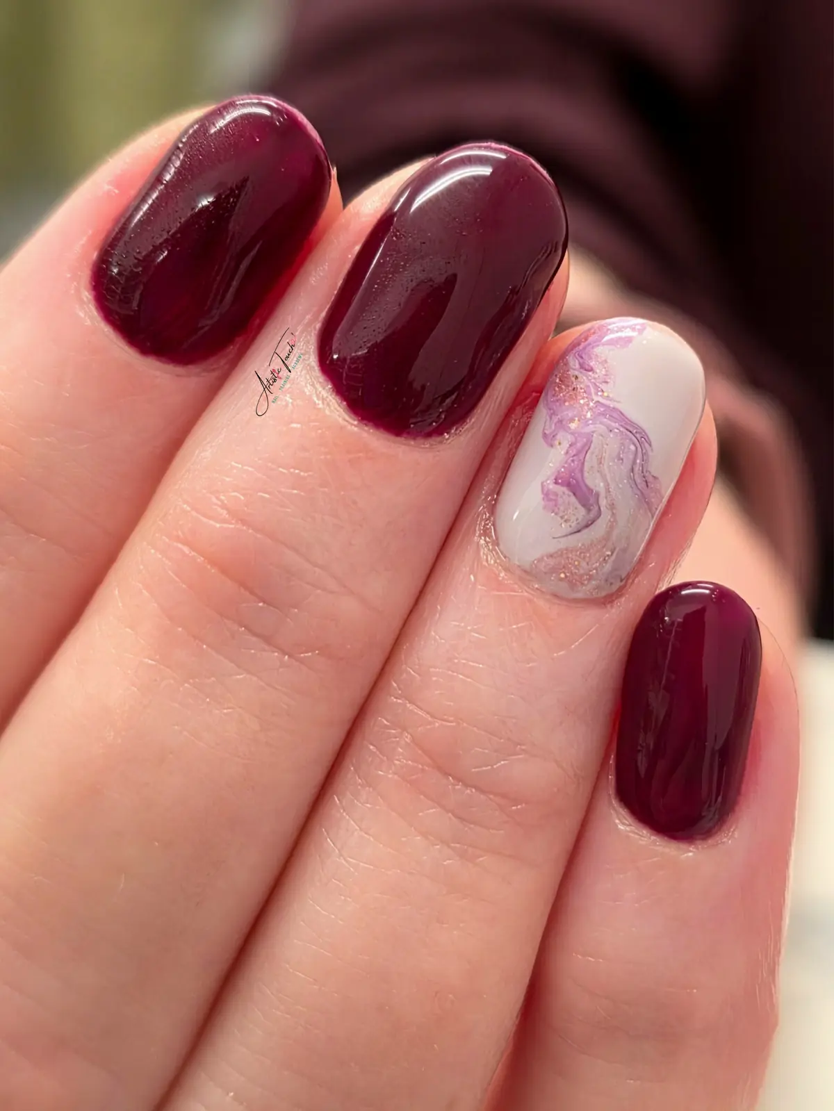 Burgundy manicure on short rounded nails with one white accent nail marbled in lilac with gold flecks