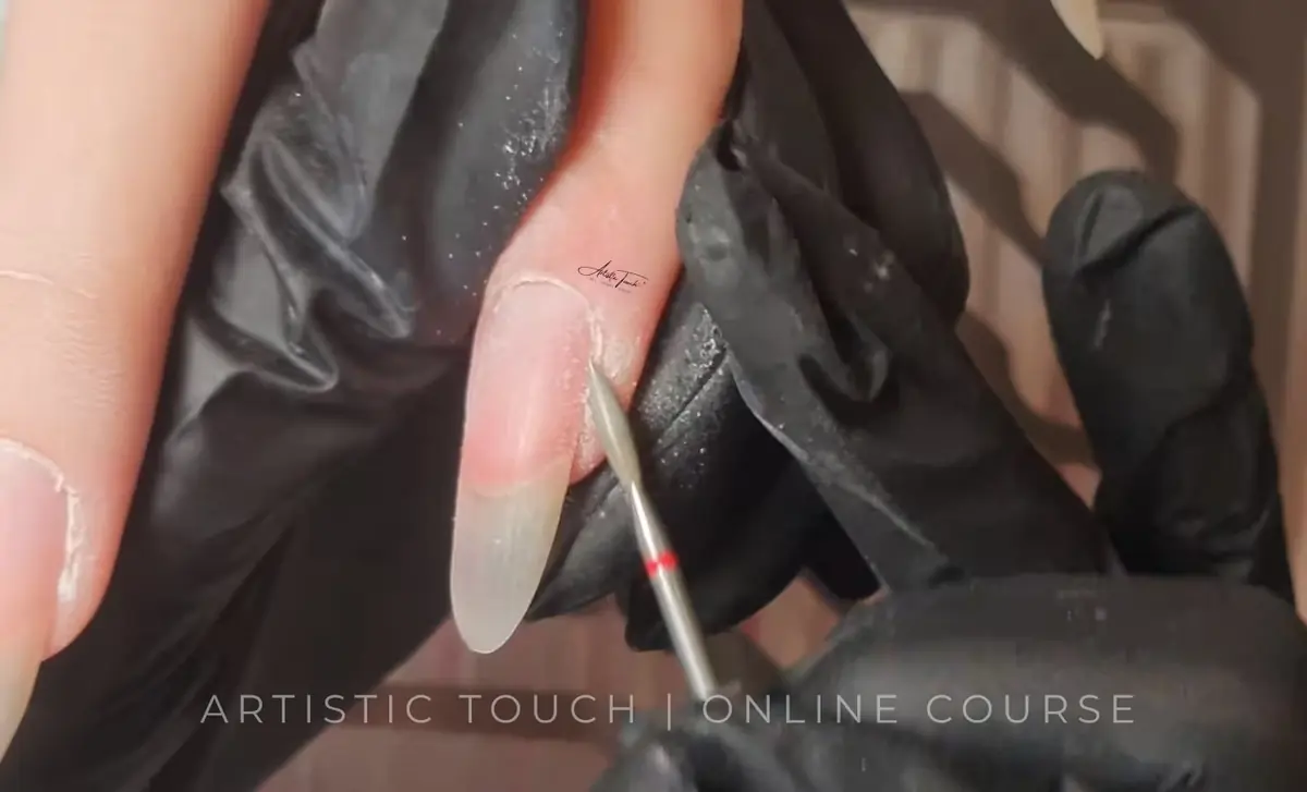 Gloved hands holding a client's finger while a fine e-file bit with a red band is positioned beside the nail, with the text Artistic Touch Online Course across the foot of the frame