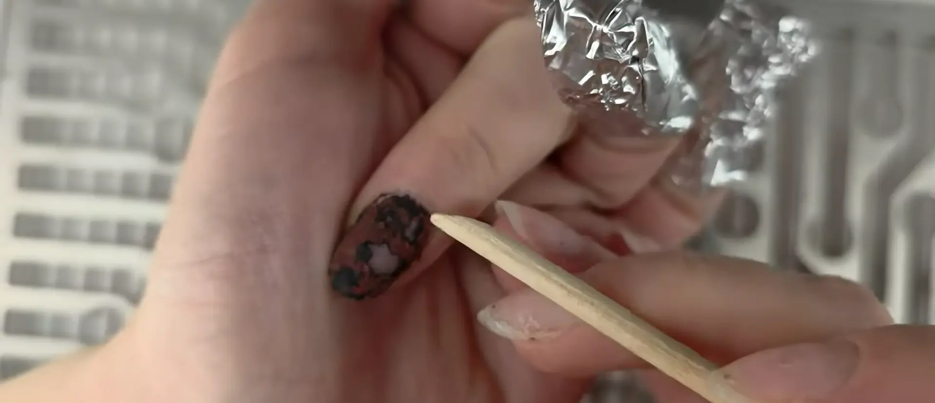 Proper technique for gently scraping softened gel polish with orange wood stick showing crumbly texture indicating correct soaking time